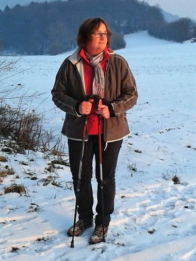 Eine Person steht auf einem verschneiten Feld und hält zwei Wanderstöcke. Im Hintergrund sind Bäume und sanfte Hügel sichtbar.