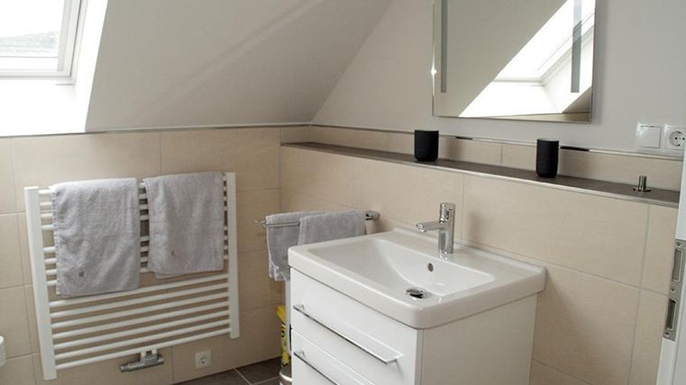 Ein modernes Badezimmer mit einer weißen Waschtischanlage und einem großen Spiegel. Handtücher hängen an einem Heizkörper und das Sonnenlicht fällt durch ein Dachfenster.