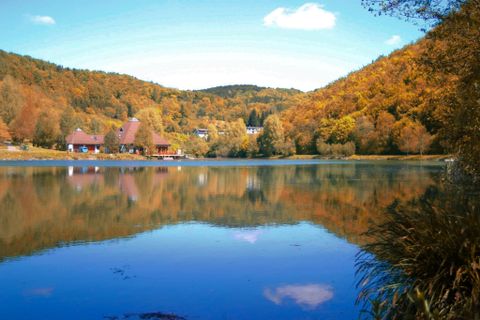 Ein ruhiger See umgeben von bunten Herbstbäumen. Im Hintergrund sind einige Häuser und ein blauer Himmel mit wenigen Wolken zu sehen.