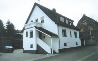 Ein modernes, weißes Wohnhaus mit einem Balkon und einer Treppe. Es befindet sich an einer ruhigen Straße, umgeben von Bäumen.