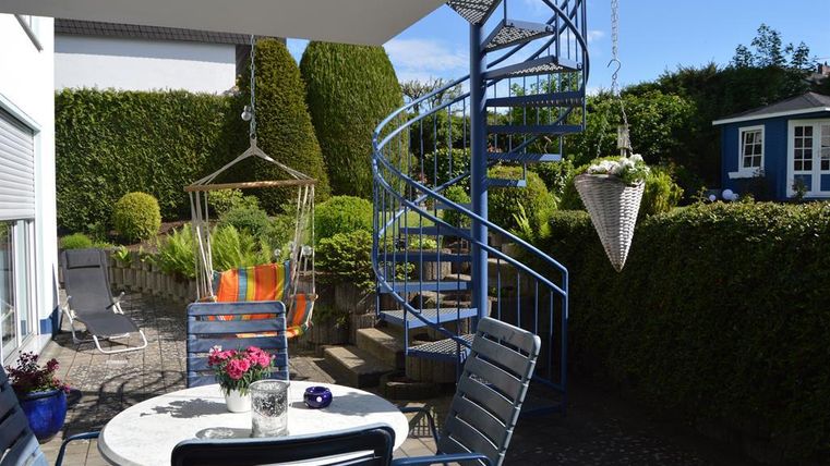 Ein schöner Patio mit einem Tisch und Stühlen. Im Hintergrund gibt es eine Wendeltreppe und üppige Gartenlandschaft.