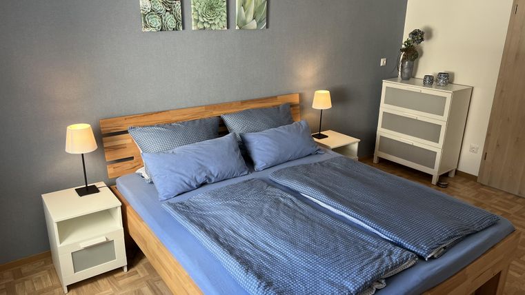 Ein gemütliches Schlafzimmer mit einem großen Bett und blauer Bettwäsche. Neben dem Bett stehen Nachttische mit Lampen und eine schlichte Kommode.