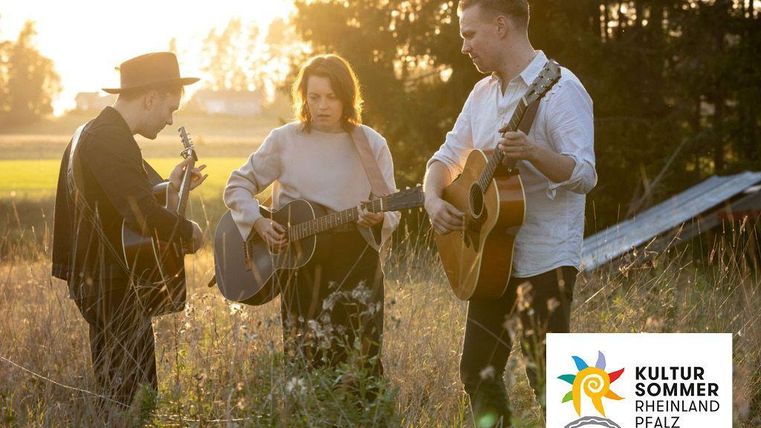 Drei Musiker spielen harmonisch Gitarre in einem Feld bei Sonnenuntergang. Die Atmosphäre ist entspannt und kreativ.