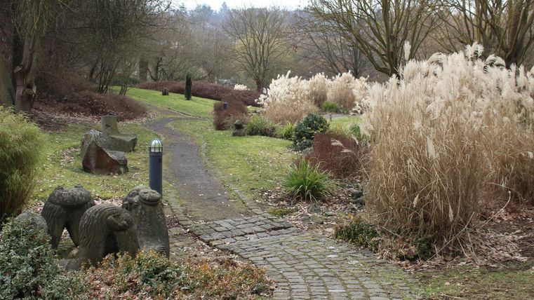 Ein malerischer Garten mit gepflastertem Weg und verschiedenen Skulpturen. Hohe Gräser und Bäume umrahmen die Szene.