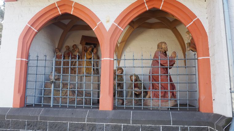 Zwei gotische Bögen mit Figuren im Inneren. Eine der Figuren zeigt eine Person in rotem Gewand, umgeben von weiteren Figuren hinter einem Gitter.