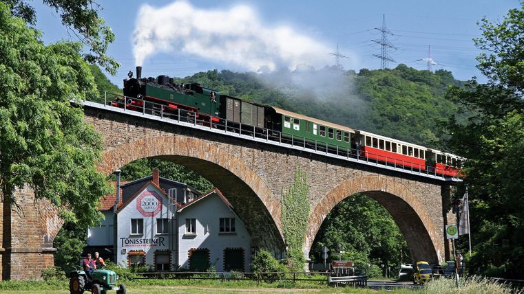 Eine dampfbetriebene Lokomotive fährt an einer Grünfläche vorbei. Familien mit Kindern beobachten den Zug aus der Nähe.