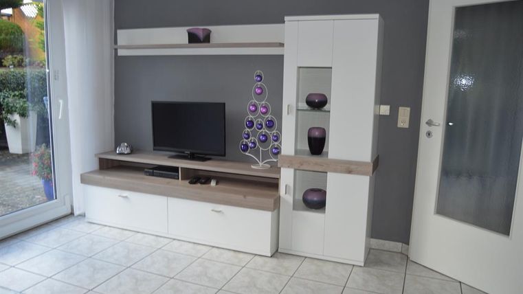 Ein modernes Wohnzimmer mit einem Fernseher und hellen Möbeln. Die Wände sind grau, und es gibt dekorative Elementen in lila.