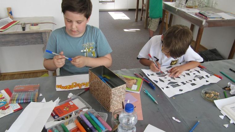 Zwei Kinder arbeiten an einem Basteltisch. Sie verwenden Buntstifte und Papiere, während im Hintergrund weitere Kinder aktiv sind.