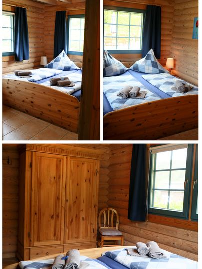 A cozy bedroom with a large double bed and wooden paneling. There is a wardrobe, a chair, and large windows with blue curtains.
