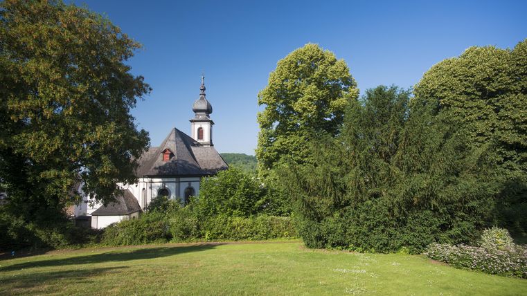 Eine schöne Landschaft mit einem grünen Rasen und Bäumen. Im Hintergrund ist eine Kirche sichtbar, unter einem klaren blauen Himmel.