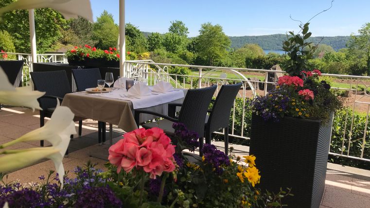 A beautiful outdoor area of a restaurant with a table and chairs. In the foreground, colorful flowers are blooming and in the background, green hills can be seen.