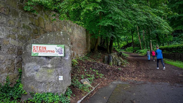 A path in the forest with a sign indicating the "Stone Experience Trail." Two people are walking along the path.