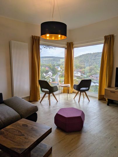Ein modernes Wohn- und Esszimmer mit zwei Stühlen und einem Tisch. Durch das große Fenster ist eine grüne Landschaft sichtbar.