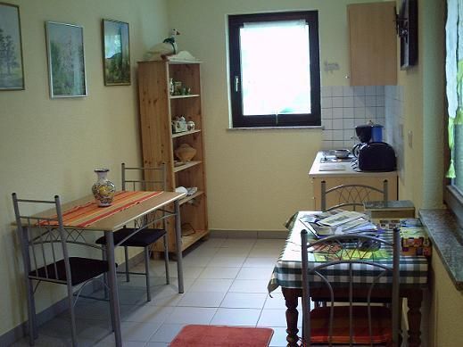 A small, cozy kitchen with a table and chairs. Pictures hang on the wall, and in the corner stands a shelf with dishes.