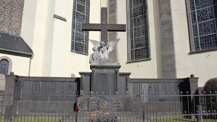 Ein Grabmal mit einem großen Kreuz und einer Statue eines Engels. Es befindet sich vor einer Kirche, umgeben von einem Eisenzaun.