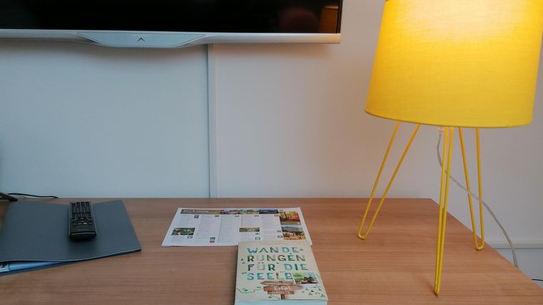 A table with a yellow lamp and a book. In the background, there is a television, and next to it lies a brochure.