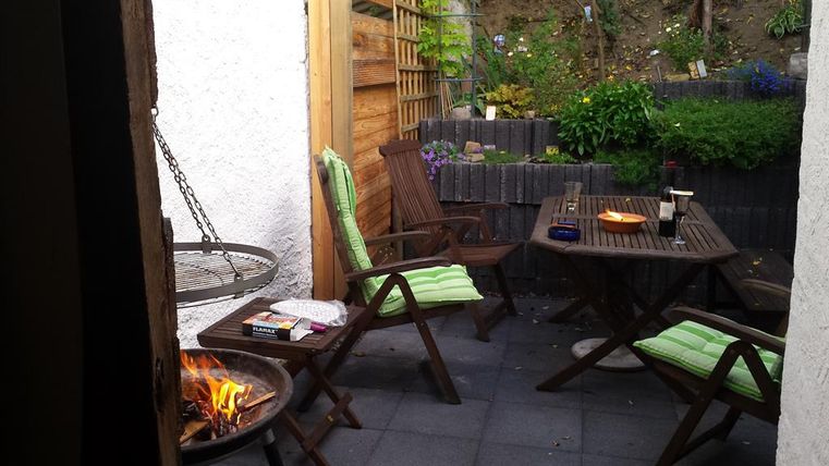 Eine gemütliche Terrasse mit Holzstühlen und einem Tisch. Im Hintergrund sind Pflanzen und ein kleiner Feuerkorb zu sehen.