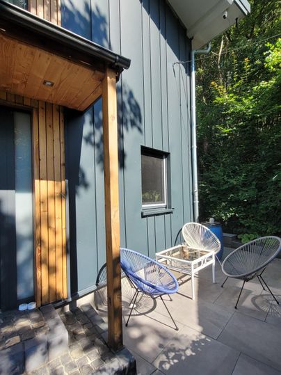 Eine moderne Terrasse mit einem blauen Holzhaus im Hintergrund. Es gibt gemütliche Stühle und viel grünes Laub.