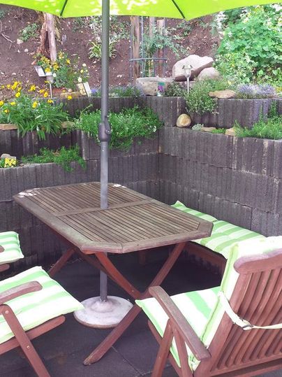 Ein gemütlicher Sitzbereich im Garten mit einem Holztisch und zwei Stühlen. Ein grüner Sonnenschirm schützt vor der Sonne und umgeben sind Pflanzen und Blumen.