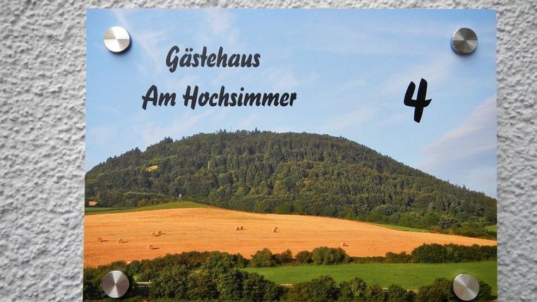 Ein Schild mit der Aufschrift "Gästehaus Am Hochsimmer" und der Zahl 4. Im Hintergrund ist eine grüne Hügellandschaft mit Feldern zu sehen.