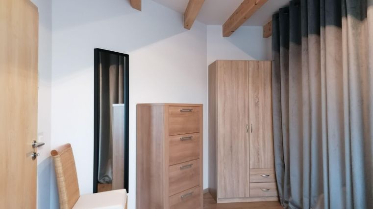A bright room with wooden paneling and a large window. In the room, there is a wardrobe, a dresser, and a chair.