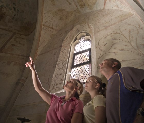 Drei Personen betrachten Fresken in einer Kirche. Eine Frau zeigt auf die Decke. Ein gotisches Fenster mit Gitter ist im Hintergrund sichtbar., © Traumpfade/Kappest