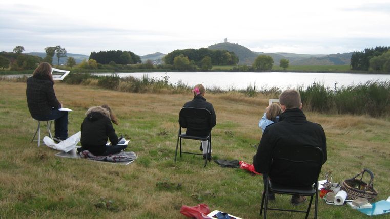 Eine Gruppe von Menschen sitzt auf Stühlen in einem Feld und malt oder zeichnet. Im Hintergrund ist ein ruhiger See und sanfte Hügel zu sehen.