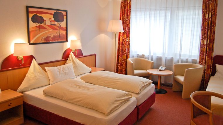 Ein gemütliches Hotelzimmer mit zwei Betten und einer großen Fensterfront. Die Wände sind freundlich dekoriert und es gibt ein paar Sitzgelegenheiten.