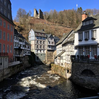 Ein malerisches Dorf mit Fachwerkhäusern entlang eines kleinen Flusses. Im Hintergrund sind Ruinen auf einem Hügel zu sehen.