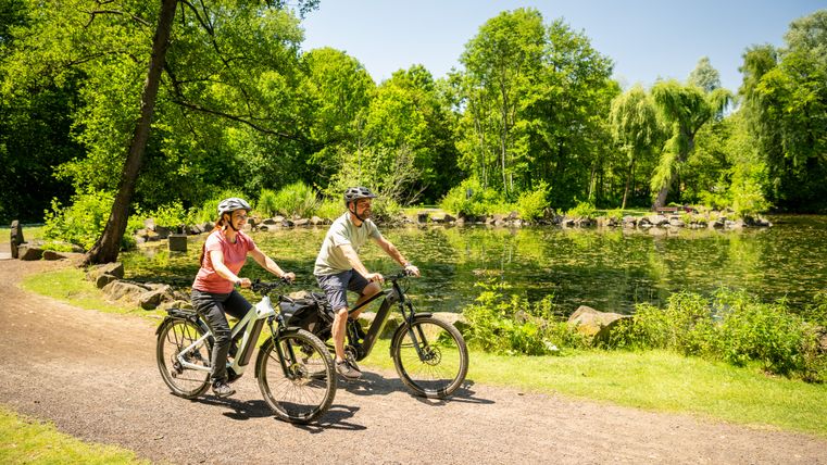 Mann und Frau auf Fahrrädern. Im Hintergrund sieht man einen kleinen Teich. Grüne Bäume rund um den Teich.