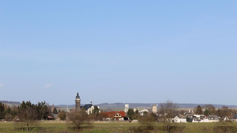 Eine malerische Landansicht mit einem kleinen Dorf und einer klaren blauen Himmel. Im Hintergrund ist ein Wasser- oder Kirchturm sichtbar.