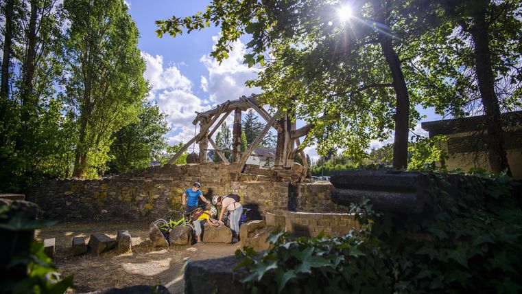 A beautiful green space with trees and a sunny sky. In the background, a stone structure can be seen, and some people are enjoying the surroundings.