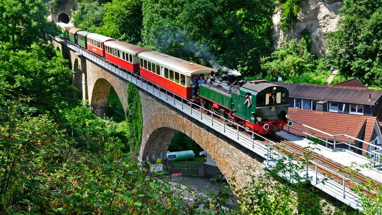Eine historische Dampflokomotive zieht bunte Waggons über eine steinerne Eisenbahnbrücke in einer grünen, bewaldeten Landschaft.