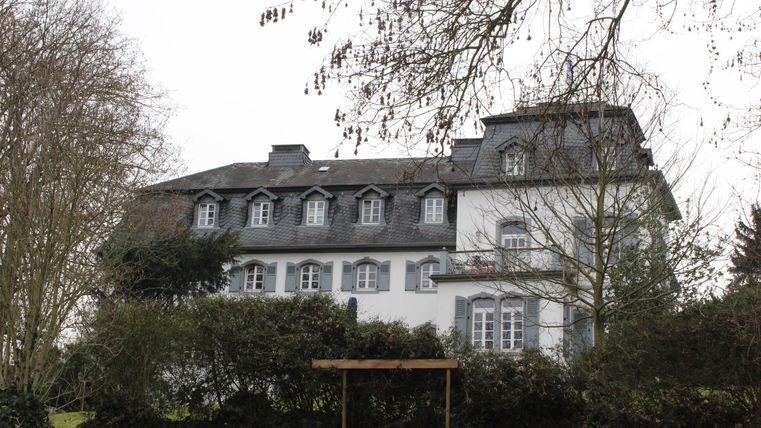 Ein historisches Gebäude mit grauem Dach und weißen Wänden. Es ist von Bäumen und Sträuchern umgeben.