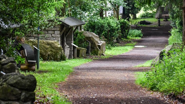 A picturesque path lined with trees and stones. The surroundings are green and inviting.