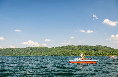 Ein sonniger Tag am See mit mehreren Fahrern auf Tretbooten. Im Hintergrund sind grüne Hügel und ein klarer blauer Himmel zu sehen.