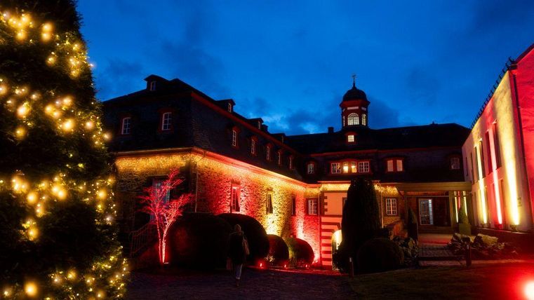 Ein historisches Gebäude beleuchtet mit roten und gelben Lichtern. Im Vordergrund steht ein geschmückter Weihnachtsbaum bei Nacht.