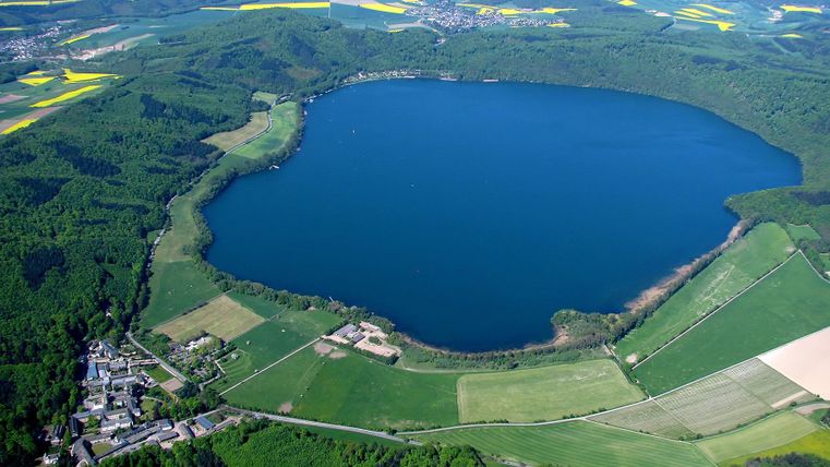 Een rustige vijver omgeven door groene velden en bossen. Op de achtergrond zijn heuvelachtige landschappen te zien.