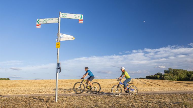 Two cyclists on a field path. In the background, there are signposts and a clear blue sky.