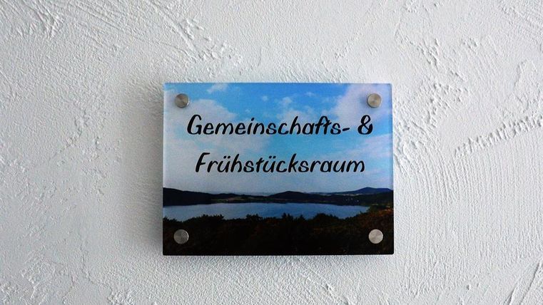 Ein Schild mit der Aufschrift „Gemeinschafts- & Frühstücksraum“ hängt an einer Wand. Im Hintergrund ist eine malerische Landschaft mit Wasser und Hügeln zu sehen.