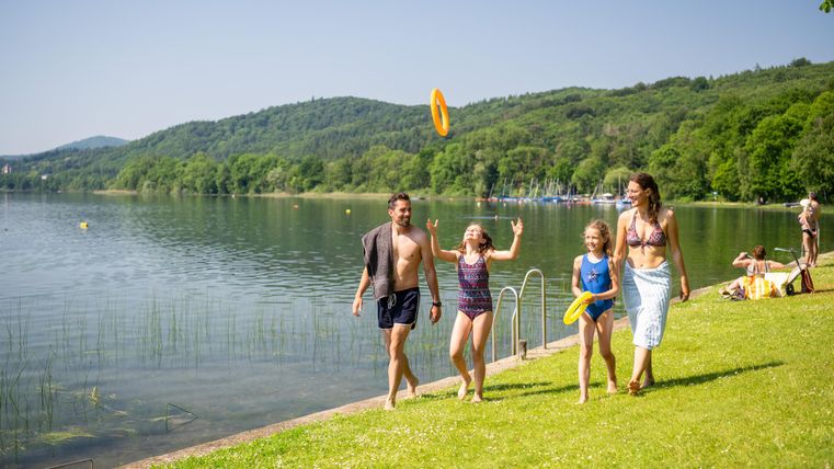 Eine Familie spielt am Ufer eines Sees. Im Hintergrund sind Bäume und ein klarer Himmel zu sehen.