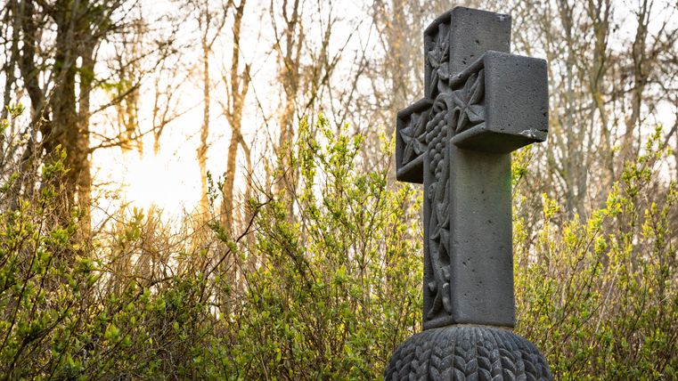 Ein schwarzes Kreuz steht in einem bewaldeten Bereich. Im Hintergrund sind sanfte Baumkonturen und das Licht der untergehenden Sonne zu sehen.