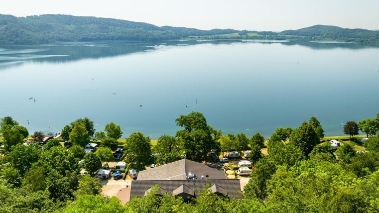Ein ruhiger See umgeben von grünen Bäumen und sanften Hügeln. Im Vordergrund sind Campingplätze und ein Gebäude sichtbar.