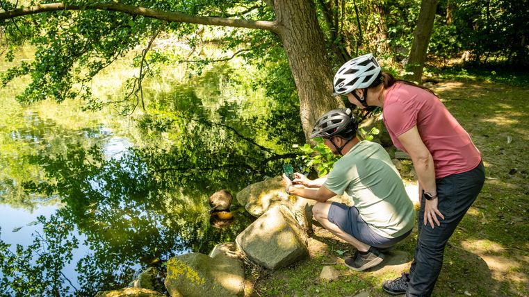 Zwei Radfahrer stehen an einem ruhigen Gewässer und schauen auf ihr Handy. Die Umgebung ist grün mit viel Licht und Bäumen.