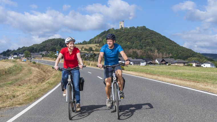 Ein Paar fährt fröhlich mit dem Fahrrad auf einer Straße. Im Hintergrund ist ein Hügel mit einer Burg zu sehen.