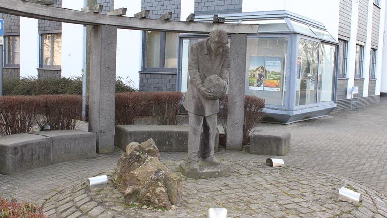 Eine Statue eines Mannes, der einen schweren Gegenstand hält, steht in einem kleinen Platz. Umgeben von Steinen und Pflanzen, wirkt die Szene ruhig und einladend.