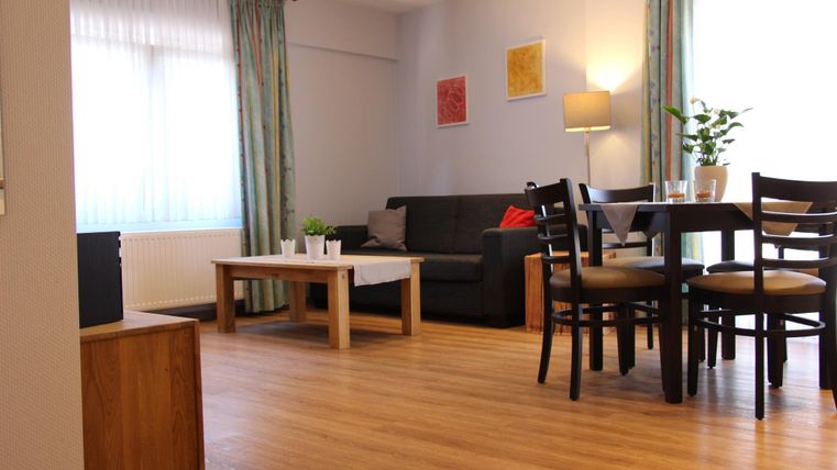 Ein gemütliches Wohnzimmer mit einem schwarzen Sofa und einem Tisch aus Holz. Lichtdurchflutete Fenster und einige Pflanzen sorgen für eine einladende Atmosphäre.