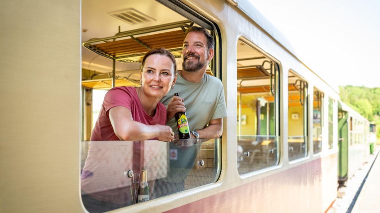 A happy couple is sitting in a colorful train, smiling out the window. They are holding a drink in their hand and enjoying the time.