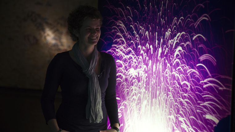 Eine lächelnde Frau steht vor einer bunten Lichtershow mit lila und weißen Strahlen. Der Hintergrund vermittelt eine aufregende und kreative Atmosphäre.