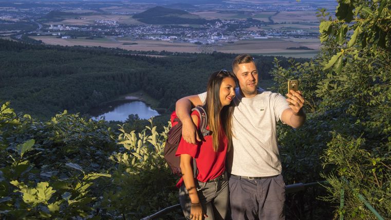 Ein Paar steht an einem Aussichtspunkt und macht ein Selfie. Im Hintergrund sind grüne Hügel und ein weites Tal zu sehen.